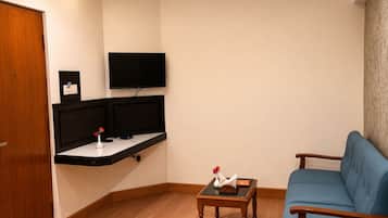 A living room with a blue sofa, a small wooden table with a vase, a flat-screen TV mounted on the wall, and a wooden door.