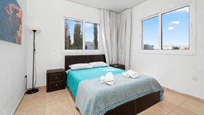 A bedroom with a bed, a nightstand, a lamp, and two windows. - Cosy 2Br Family Apartment - Mythical Sands Resort (Paralimni)