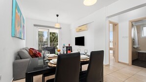 A dining area with a table set for four, a sofa, a TV mounted on the wall, and a window providing a view of the outdoors. - Cosy 2Br Family Apartment - Mythical Sands Resort (Paralimni)