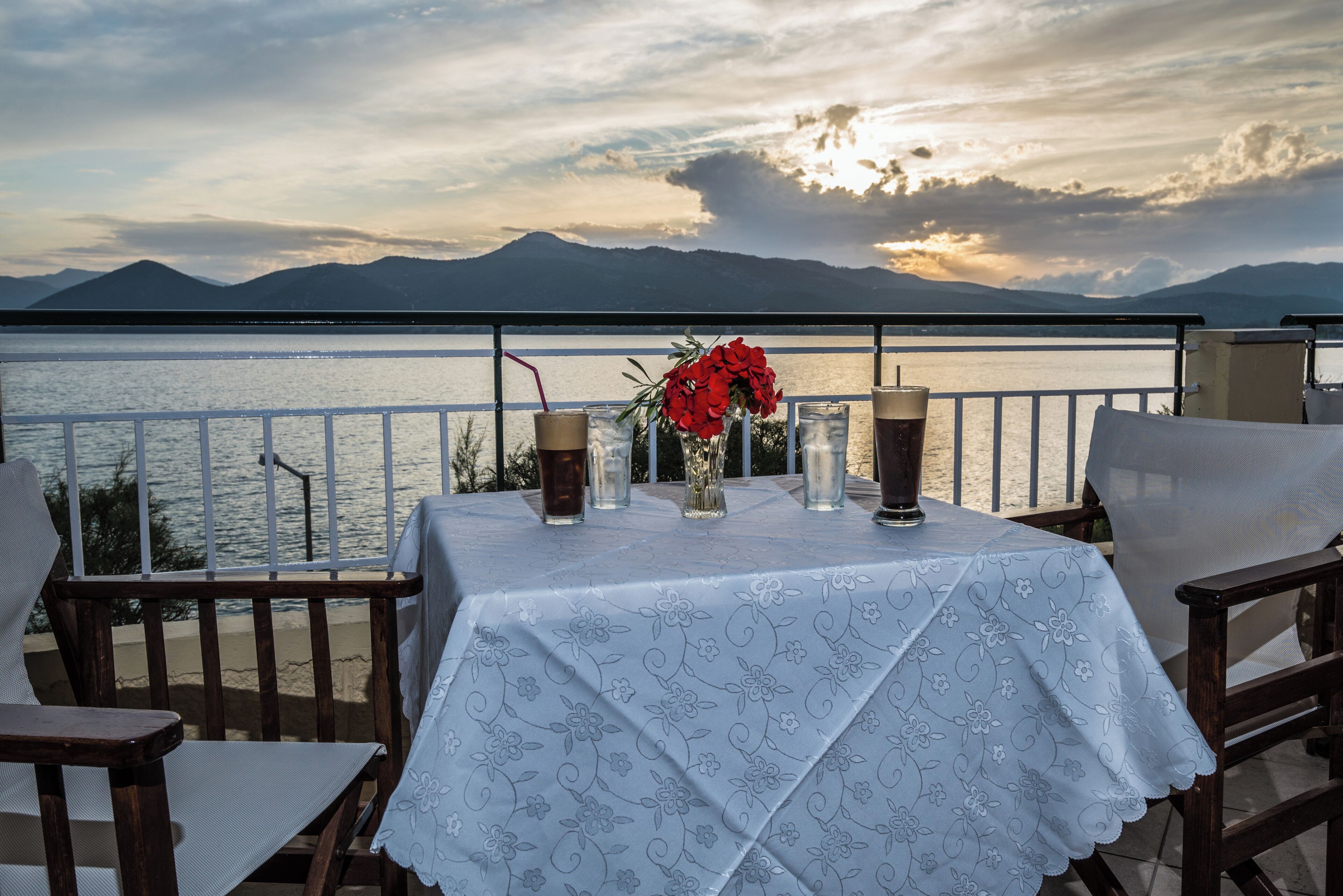 A table with a floral tablecloth, two glasses, a vase with red flowers, and a view of mountains and water.