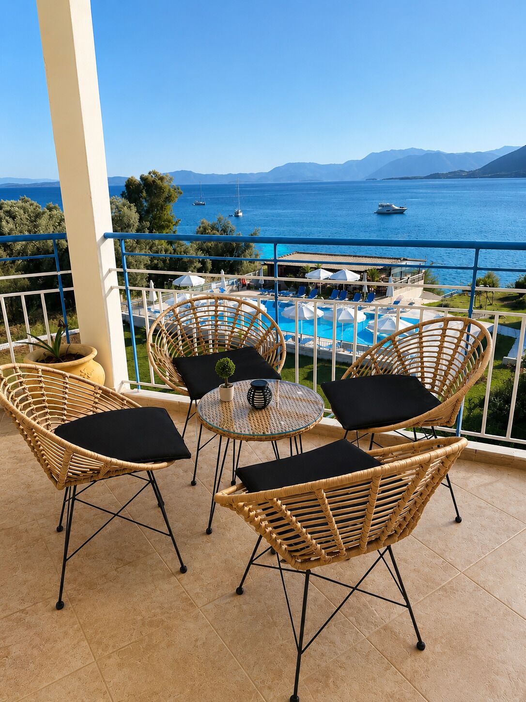 A balcony with wicker chairs and a small table, overlooking a pool and the sea.