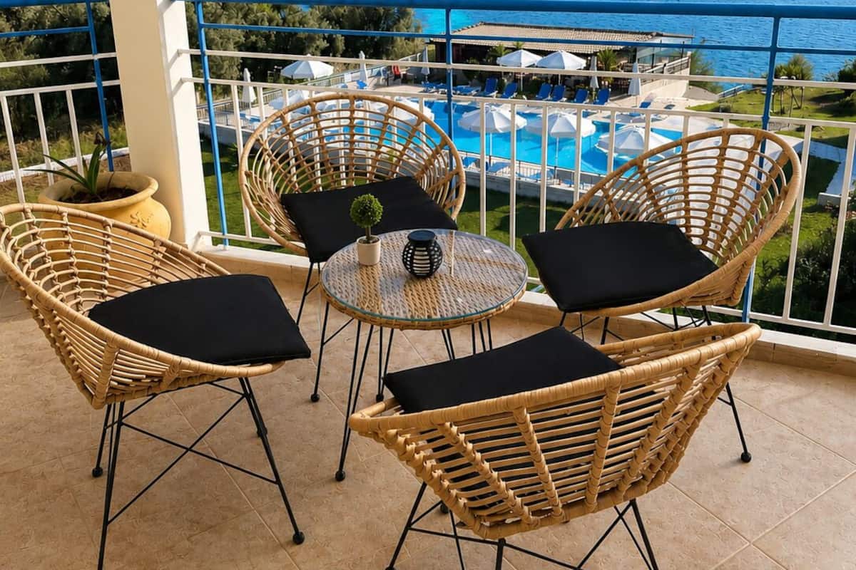 A balcony with wicker chairs and a small table, overlooking a pool and the sea.