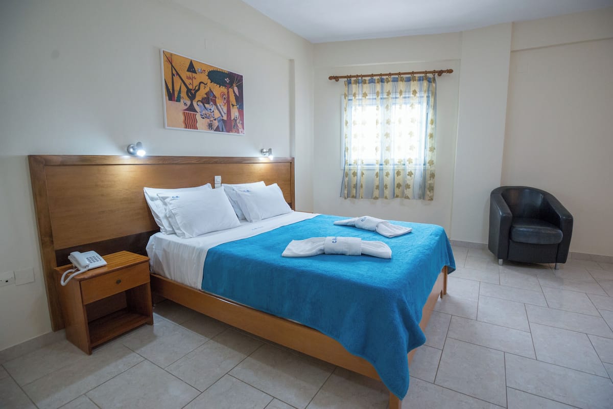 A hotel room with a bed, a nightstand with a phone, a chair, a window with curtains, and a painting on the wall.