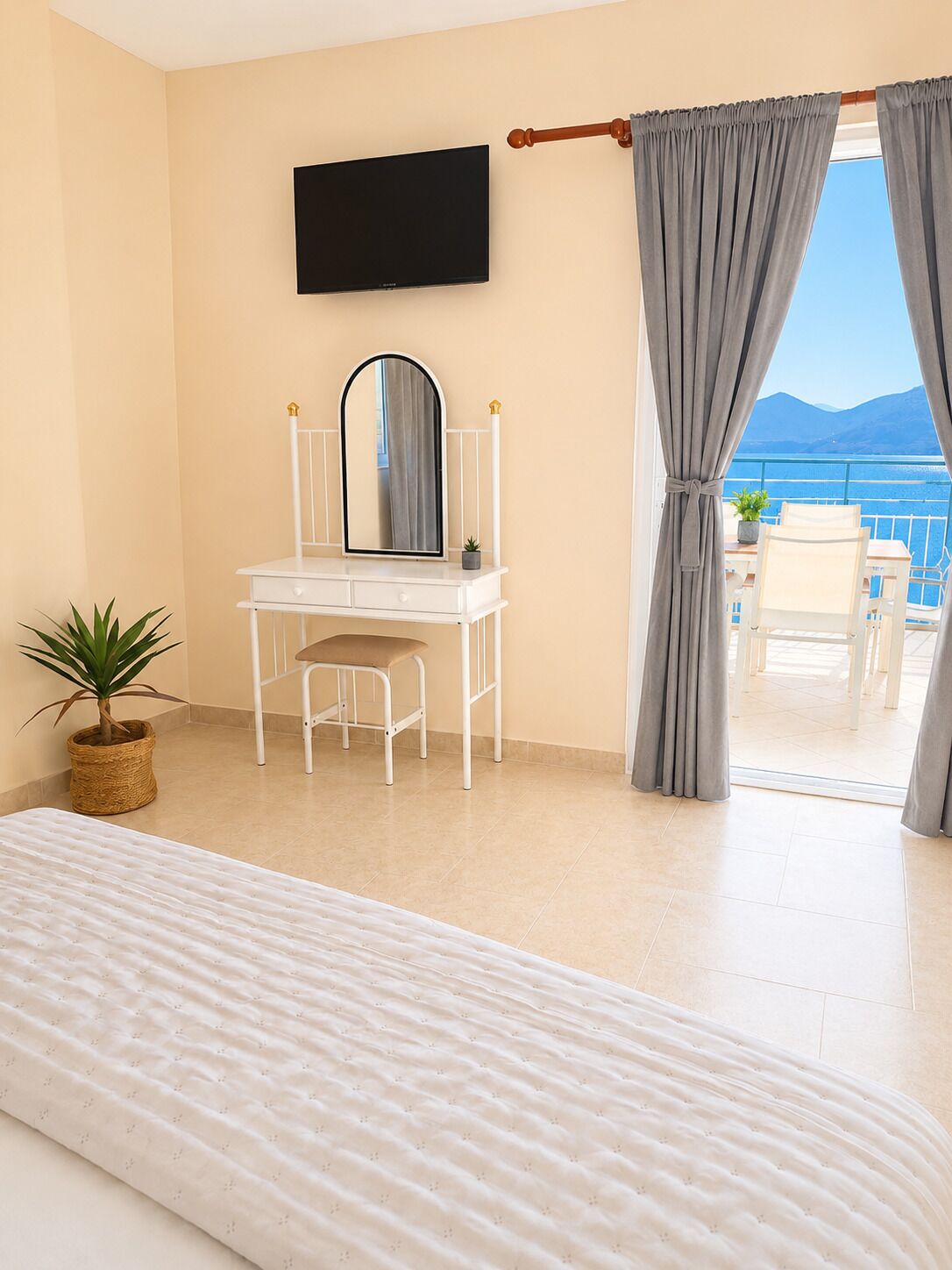 A bedroom with a bed, a desk with a mirror, a potted plant, and a view of the sea.