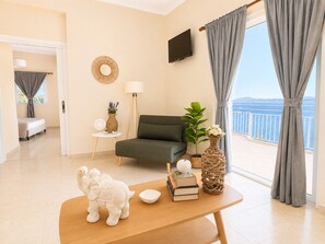 A modern living room with a sofa, a table with books and decorative items, a TV, and a balcony with curtains.