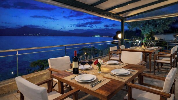 A dining area with tables set for two, overlooking a body of water with a mountainous backdrop. - Cliff Bay Hotel by GHH (Aktio-Vonitsa)