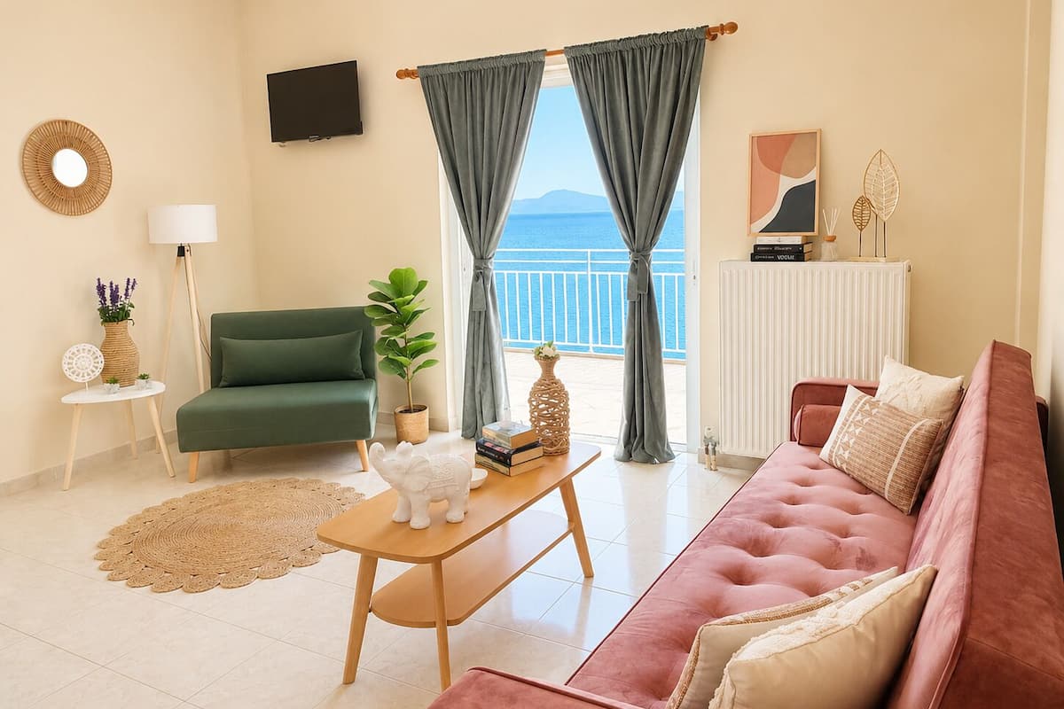 A living room with a green sofa, a pink sofa, a wooden coffee table, a small white table with a lamp, a potted plant, and a view of the sea through large windows.
