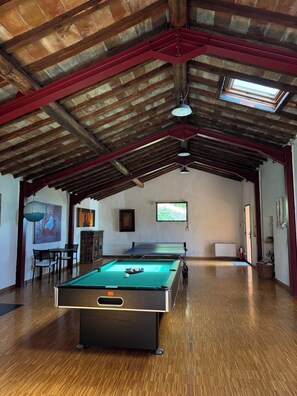Uma mesa de bilhar em um ambiente com piso de madeira, teto alto com vigas aparentes e uma janela que oferece luz natural.