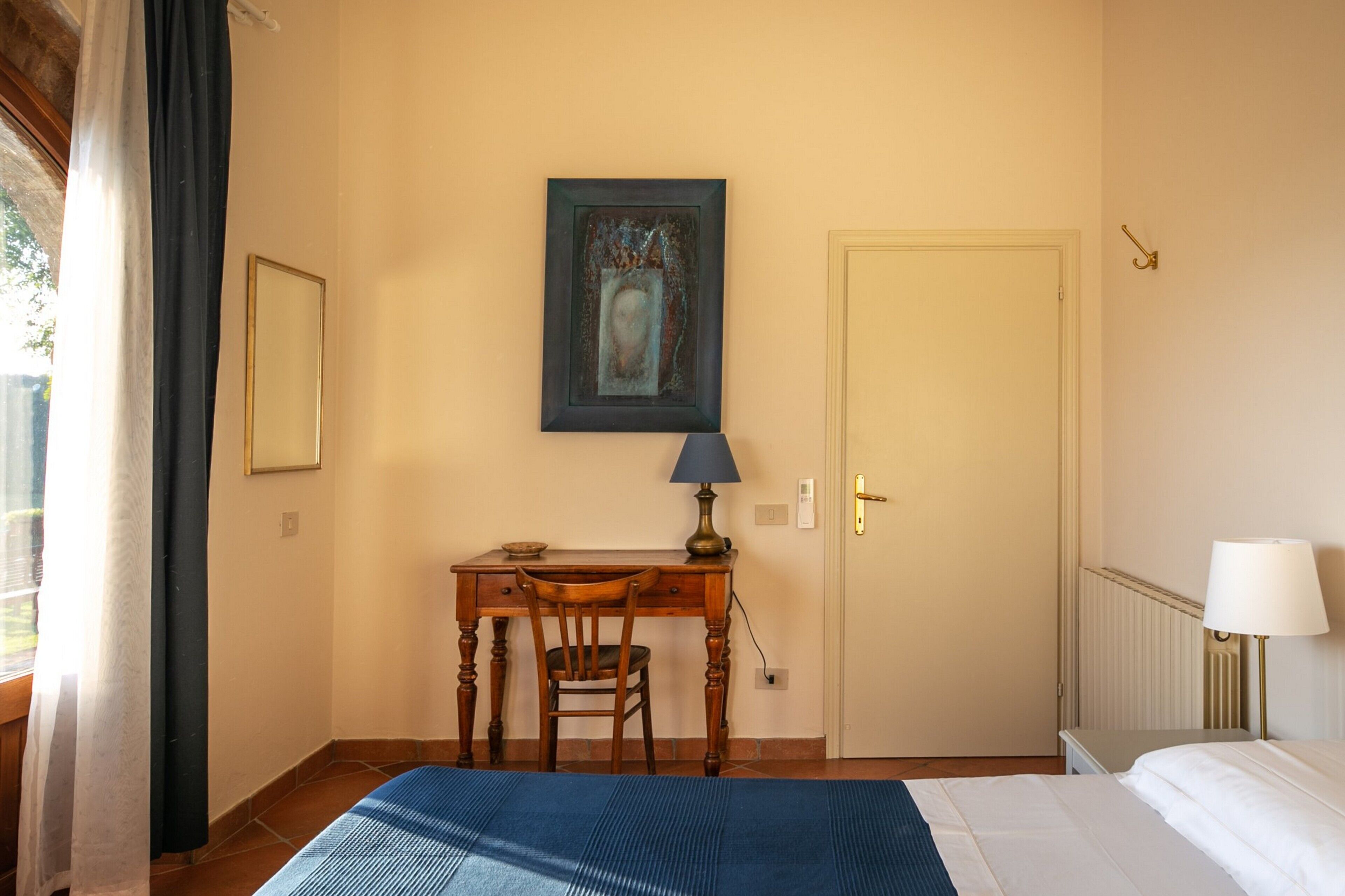 A bedroom with a bed, a desk with a chair, a lamp, a painting on the wall, and a door.