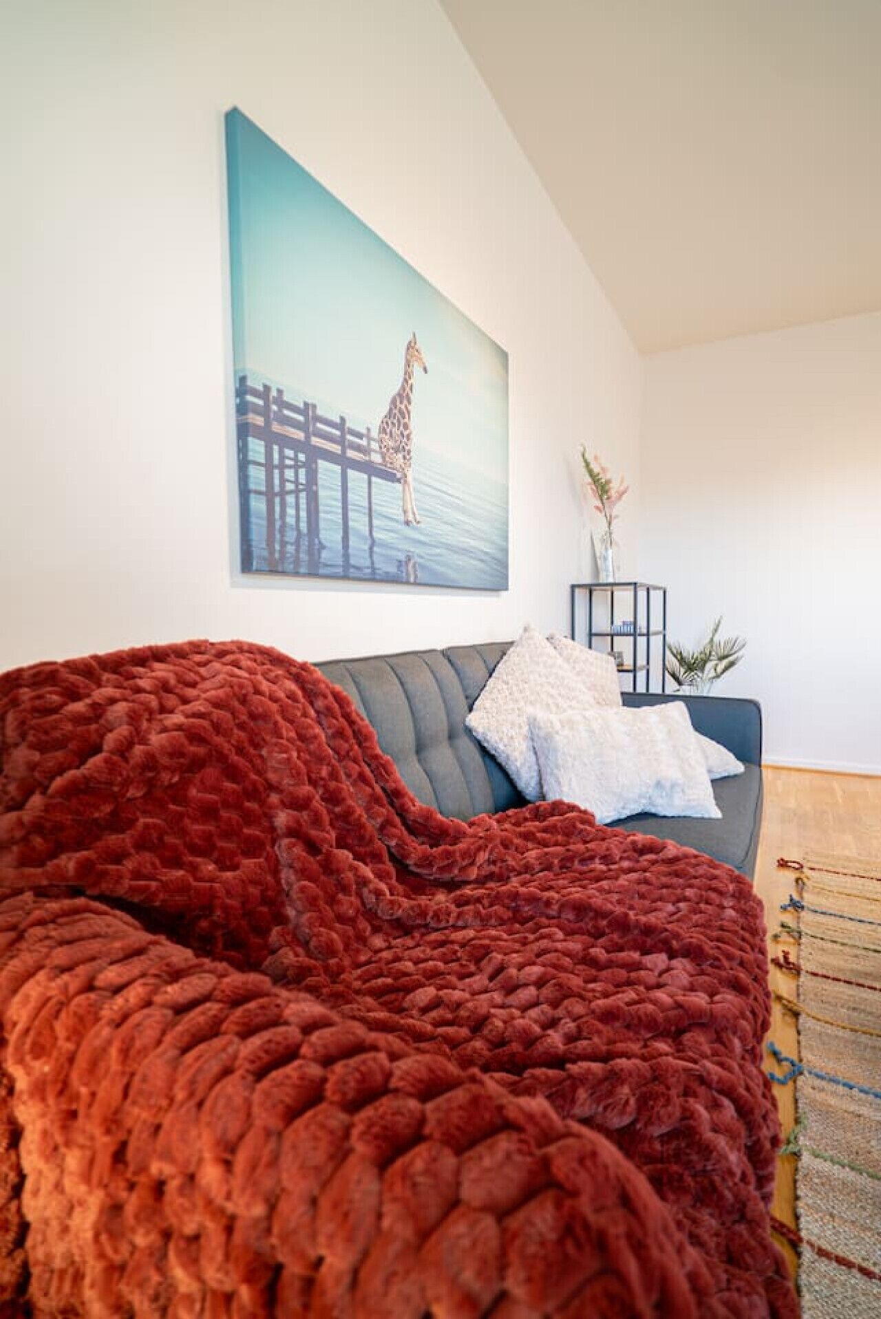 Een moderne woonkamer met een grijze bank, een rood gebreid plaid, een wit kussen, een schilderij van een giraffe op een steiger, en een kleine plant op een bijzettafeltje.