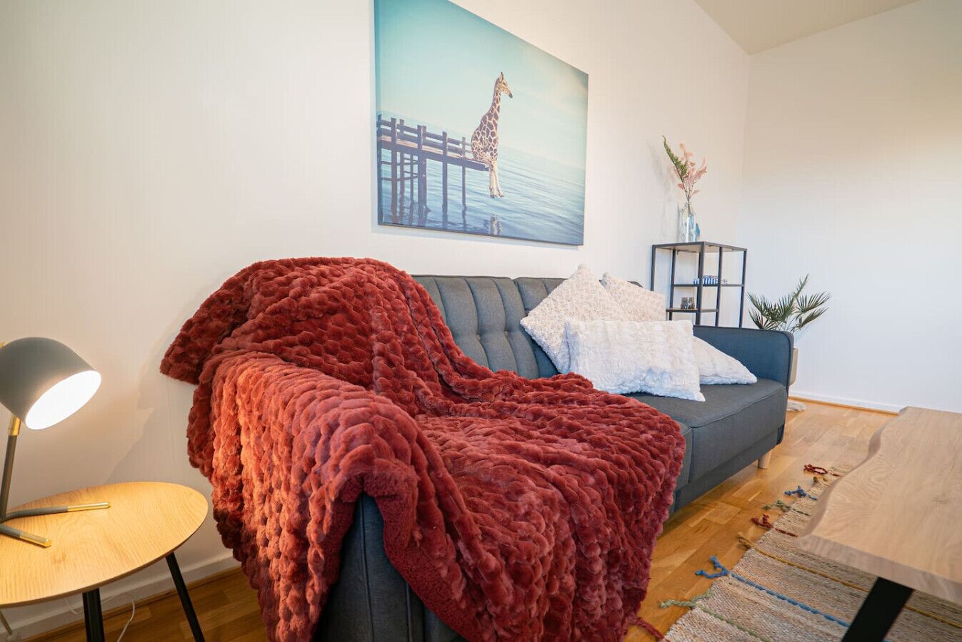 A living room with a grey sofa, a red blanket, a lamp, and a painting of a giraffe on a pier.