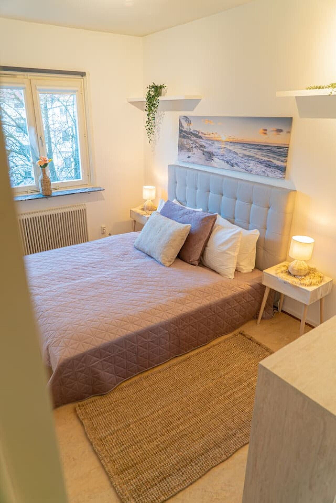 Ein Schlafzimmer mit Bett, Nachttischen, einem Fenster, einem Bild und einer Heizung.