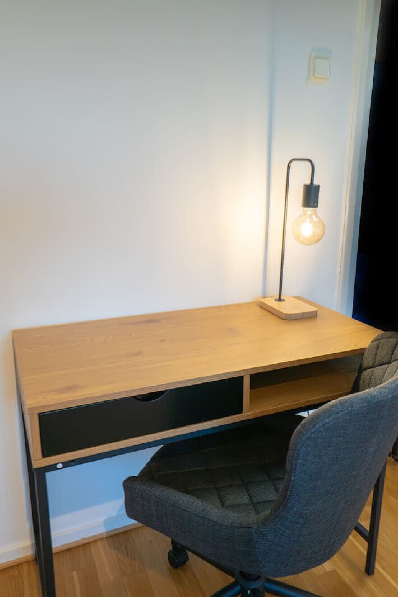 Una scrivania in legno con un cassetto nero, una sedia e una lampada a parete.