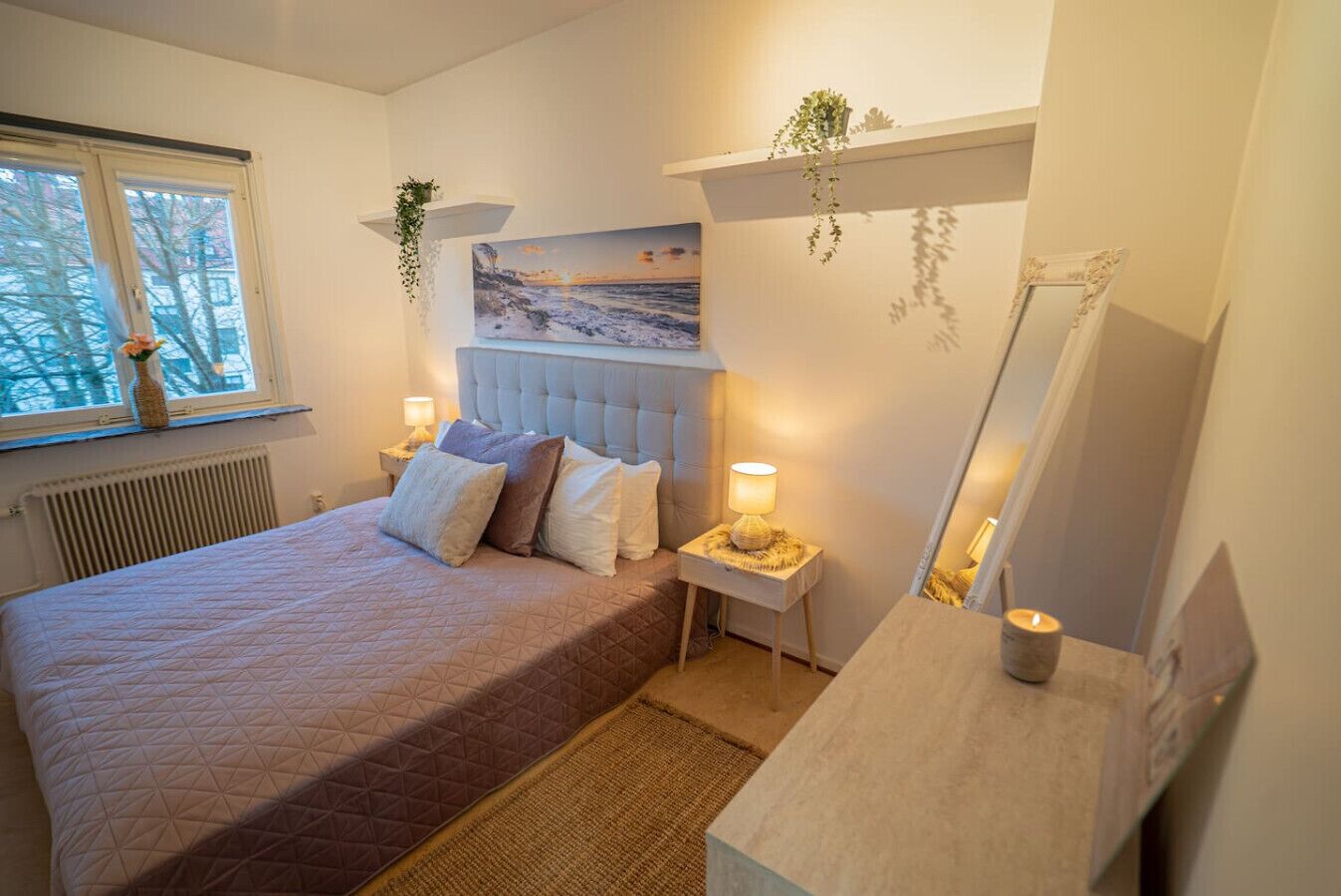 A bedroom with a bed, bedside lamps, a nightstand, a mirror, a window, and a painting on the wall.