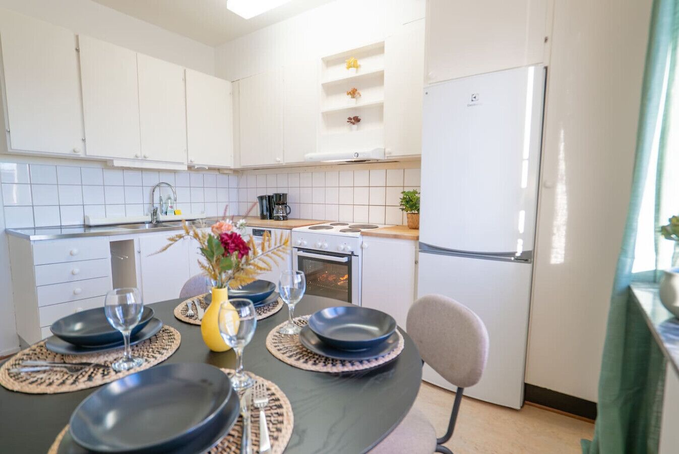 Een moderne keuken met witte kastjes, een eettafel gedekt met borden en wijnglazen, en een koelkast.
