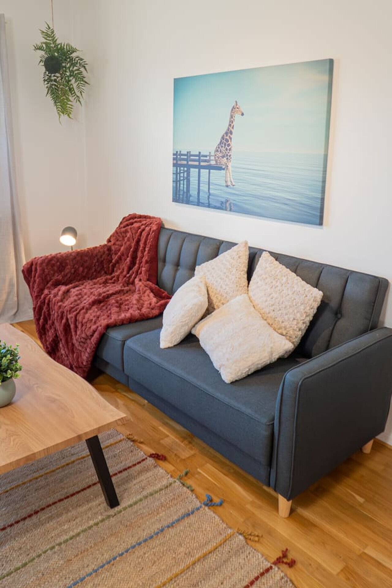 Un salon avec un canapé gris foncé, un plaid rouge et un tableau représentant une girafe accroché au mur.