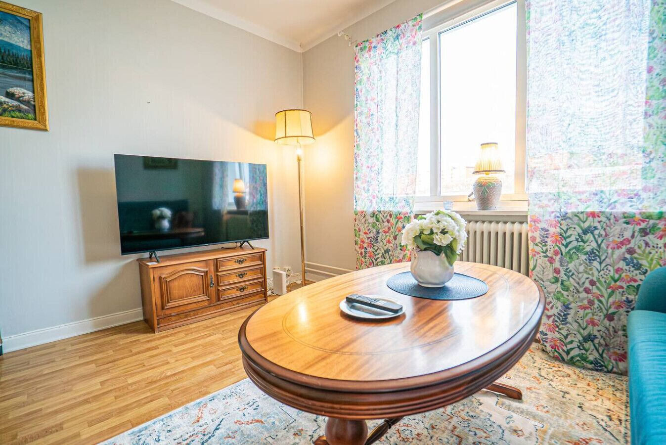 A living room with a wooden table, a television, a floor lamp, and a window with floral curtains.