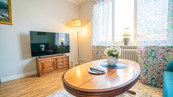 A living room with a wooden table, a television, a floor lamp, and a window with floral curtains.