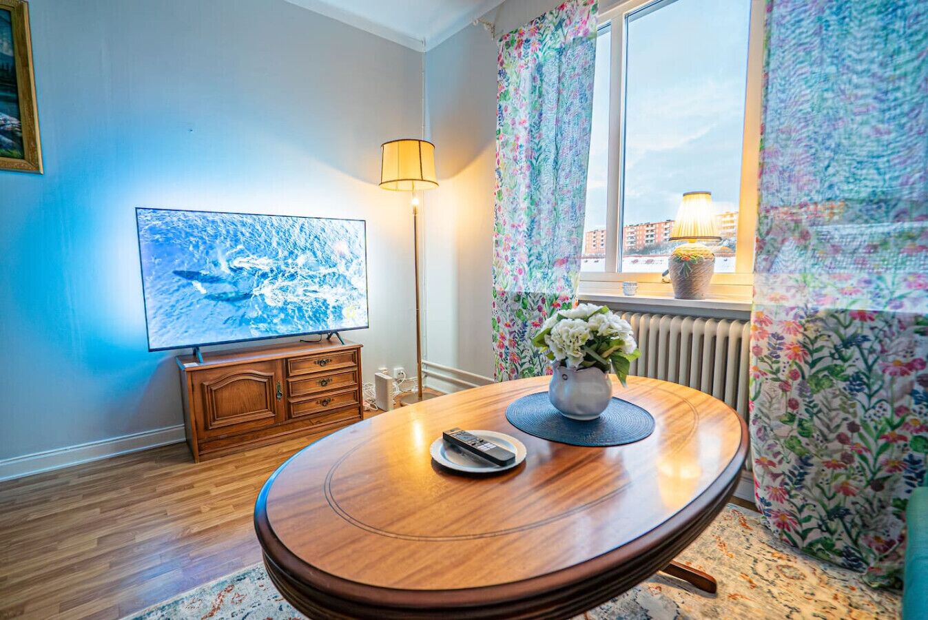 Een kamer met een houten eettafel, een televisie aan de muur, een lamp en een raam met gordijnen.