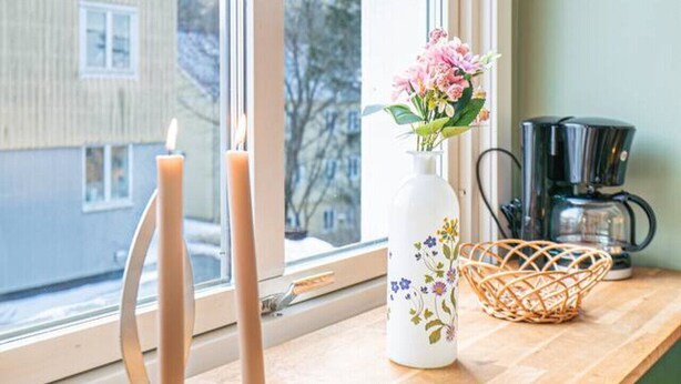 Una finestra con vista su un edificio, un vaso con fiori, un portacandele e una macchina da caffè su un ripiano di legno.