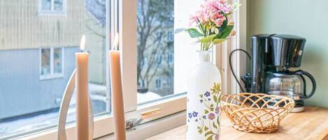 Une fenêtre donnant sur un immeuble, un vase rempli de fleurs, un chandelier et une machine à café, le tout posé sur une étagère en bois.