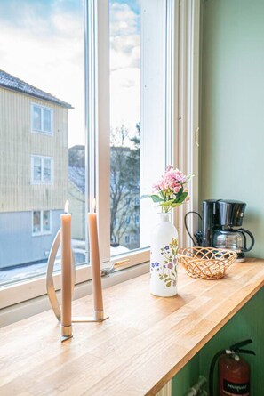 Una ventana con vista a un edificio, un florero con flores, un candelabro y una cafetera sobre un repisa de madera.
