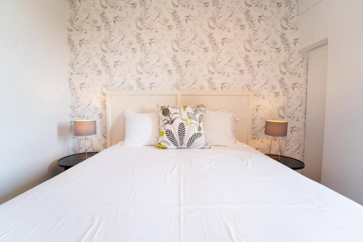 A neatly made bed with a white bedspread, two bedside lamps, and a floral wallpaper pattern.