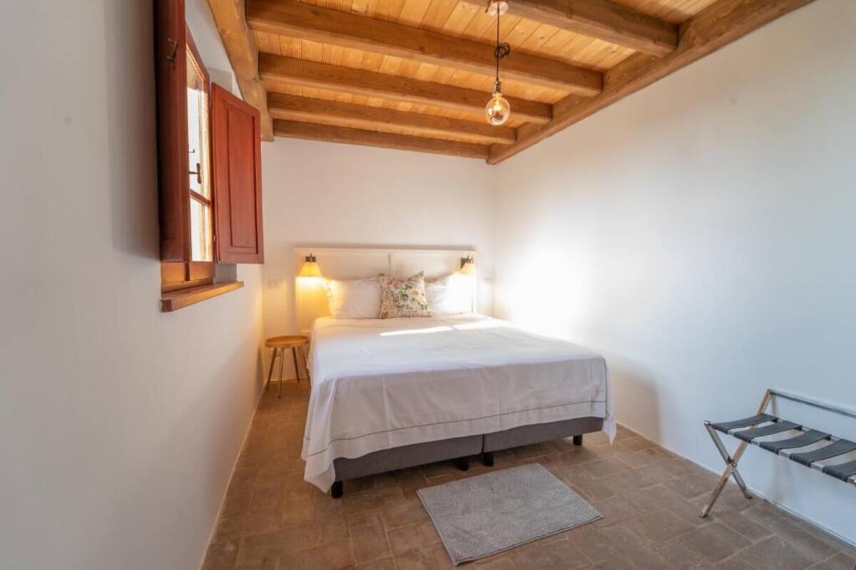 A bedroom with a wooden ceiling, a bed with a white bedspread, two bedside lamps, a nightstand, and a small window with a wooden shutter.