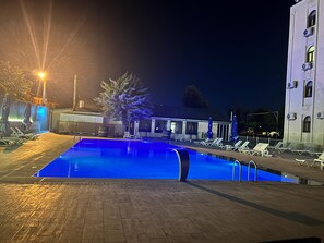 Una piscina con sillones de descanso y sombrillas, un edificio con varias ventanas y un árbol.