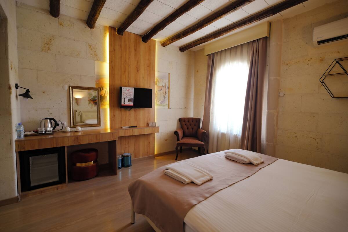 A hotel room with a wooden ceiling, a bed with a beige bedspread, a chair, a small desk with a mirror, and a wall-mounted TV.