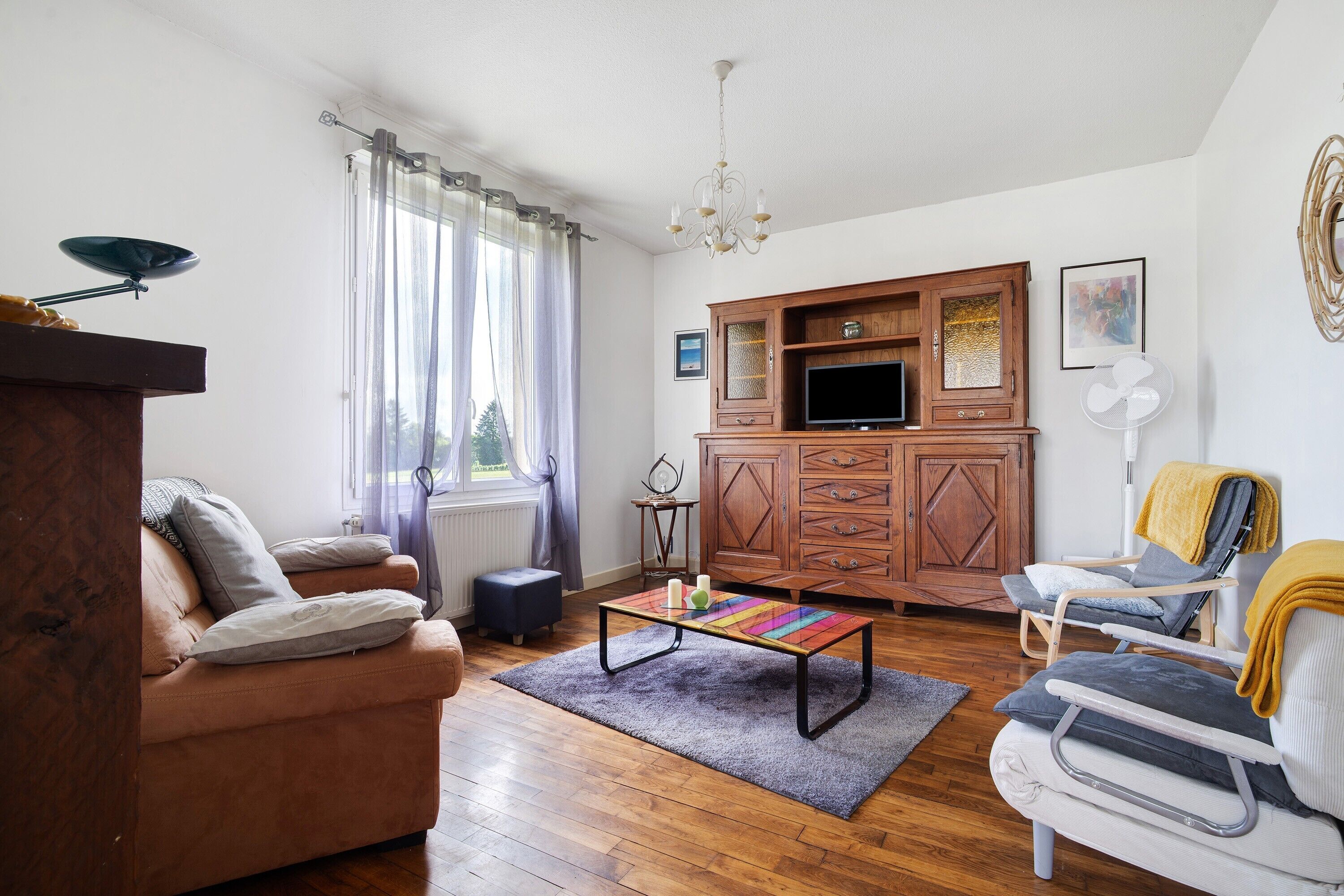 A living room with a sofa, armchair, wooden cabinet, television, and a colorful table.