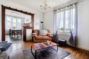 Een woonkamer met een houten vloer, een kleurrijk bijzettafeltje, een bank, een eethoek met een tafel en stoelen, en een raam met gordijnen.