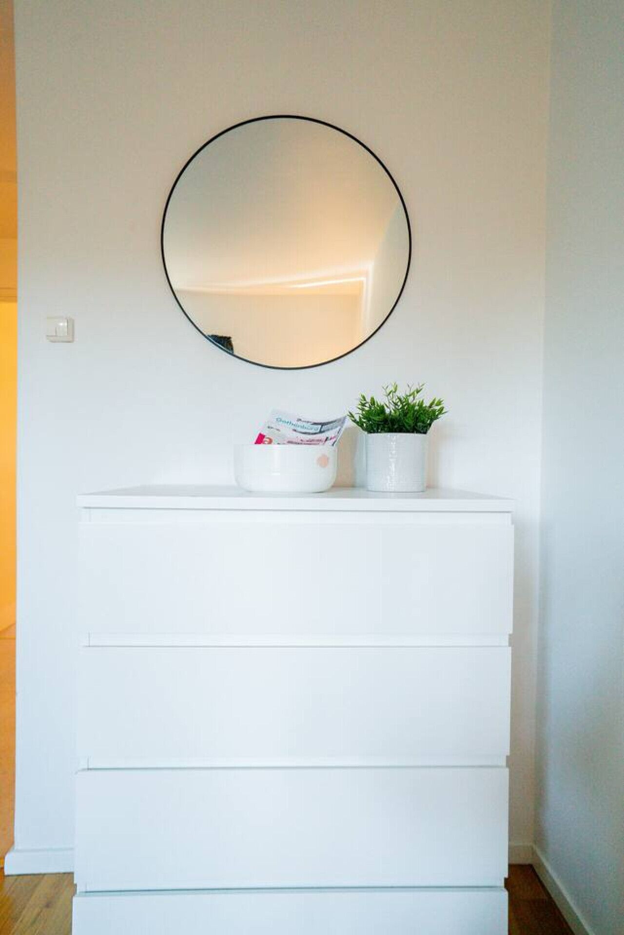 Une commode blanche avec un miroir rond au-dessus, une petite plante en pot et un bol contenant divers objets sur l’étagère supérieure.
