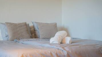 Un letto con una coperta bianca e cuscini, una parete spoglia e una finestra.
