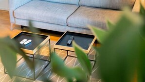 A light grey sofa with a remote control on a glass coffee table, a painting on the wall, and a plant in the foreground. - Central Studio, Near City & Tram | Brämaregården (Gothenburg)
