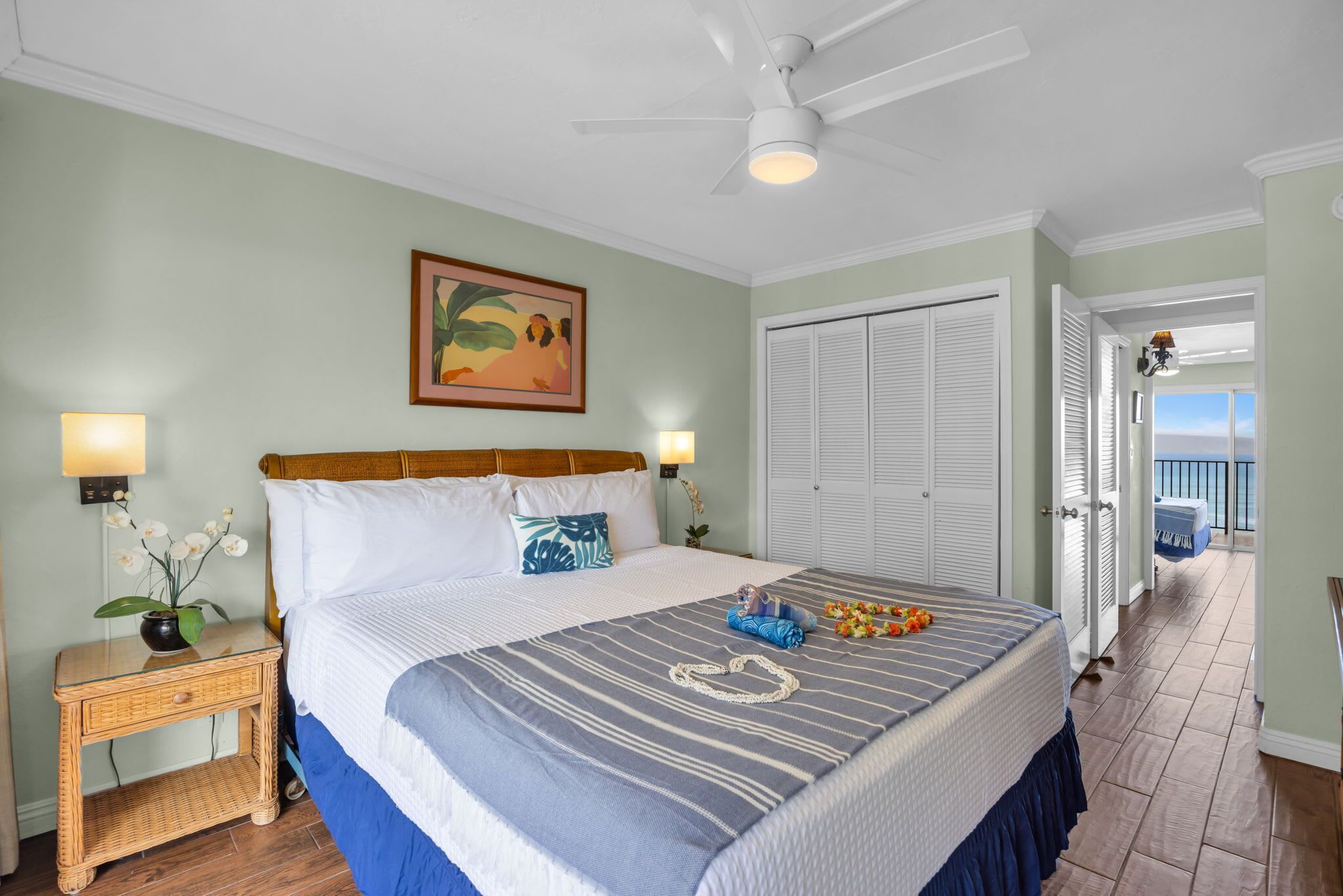 A bedroom with a bed, bedside lamps, a ceiling fan, a painting, a door leading to a balcony, and a nightstand with a vase of flowers.