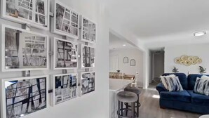 A modern living room with a blue sofa, a dining area with a white table, and a hallway with framed pictures on the wall.