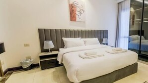A modern hotel room with a large bed, a bedside table with a lamp, a wall-mounted artwork, and a large window. - Luxury 2BR • Porto New Cairo • Pool & 3 Baths (New Cairo 1)