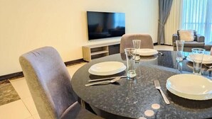 A dining area with a round table set for two, a flat-screen TV on the wall, and a sofa in the background.