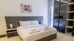 A modern hotel room with a large bed, a bedside table, a nightstand, a painting on the wall, and a view of another room through a glass door. - Luxury 2BR • Porto New Cairo • Pool & 3 Baths (New Cairo 1)