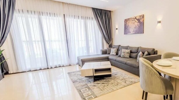 A modern living room with a sofa, a coffee table, a dining table with chairs, and large windows with curtains. - Luxury 2BR • Porto New Cairo • Pool & 3 Baths (New Cairo 1)