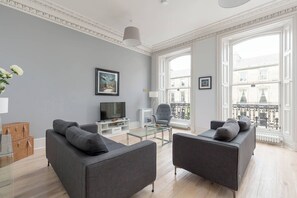 A modern living room with two grey sofas, a glass coffee table, a flat-screen TV, and a balcony with a view of buildings. - Tay Apartment (Edinburgh)