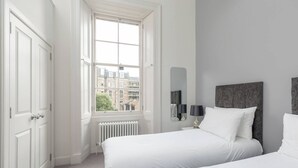 A bedroom with two beds, a radiator, a window with a view of buildings, and a mirror on the wall. - Tay Apartment (Edinburgh)