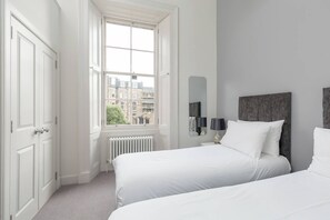 A bedroom with two beds, a radiator, a window with a view of buildings, and a mirror on the wall. - Tay Apartment (Edinburgh)