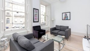 A modern living room with two grey sofas, a black armchair, and a glass coffee table. There are large windows with white frames, and artwork is displayed on the walls. - Tummel Apartment (Edinburgh)