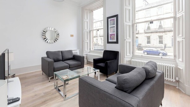 A modern living room with two grey sofas, a glass coffee table, a black armchair, a round mirror, and large windows with a view of buildings and a car. - Tummel Apartment (Edinburgh)