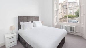 A bedroom with a bed, a nightstand, a lamp, and a window with a view of buildings and a car. - Tummel Apartment (Edinburgh)