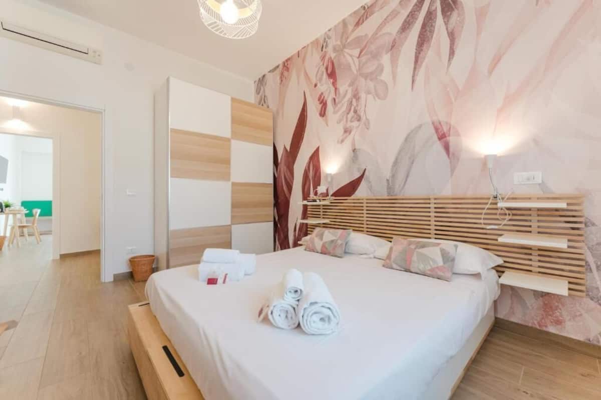 A hotel room with a bed, a wooden headboard, a wall with a floral pattern, and a small table with towels.