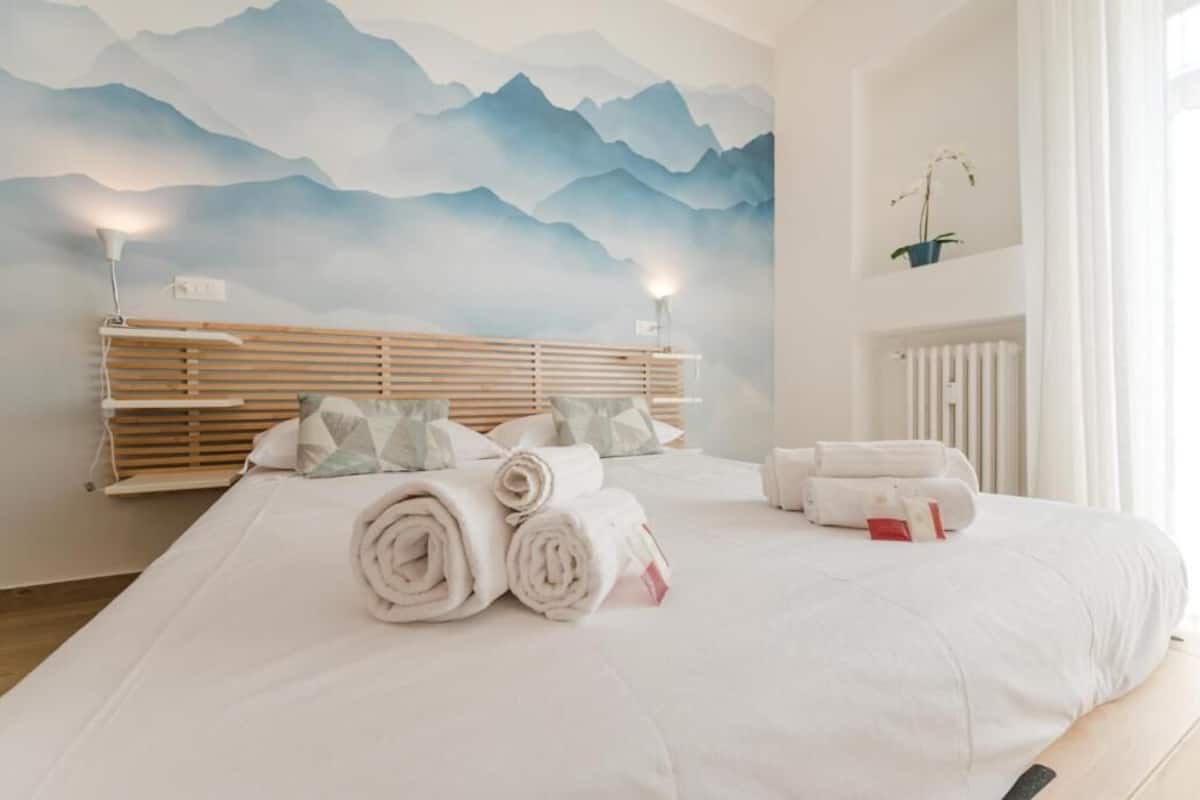 A hotel room with a bed, a headboard, a wall mural, a radiator, and a potted plant.