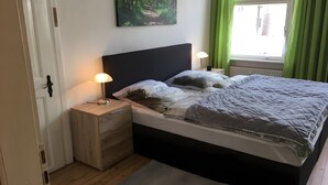 A bedroom with a bed, bedside tables, a nightstand, a lamp, a painting on the wall, and a window with curtains. - Holiday Apartment for 2 Guests (63 m²) in Zittau (Zittau)